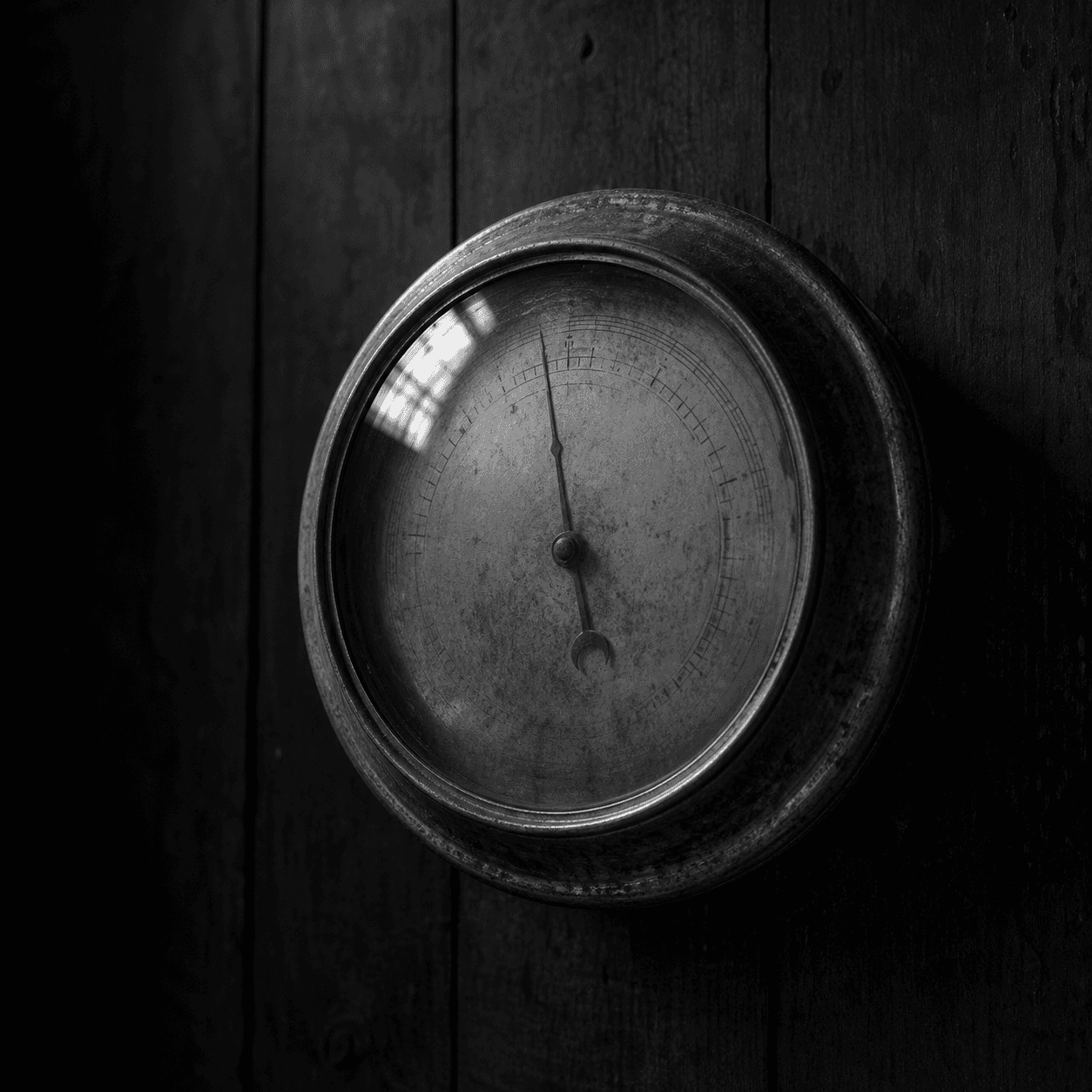 A single old brass-and-glass aneroid barometer mounted on dark wooden wall paneling. Soft dim light from above-left; the dial face is partially in shadow with a faint reflection on the curved glass.