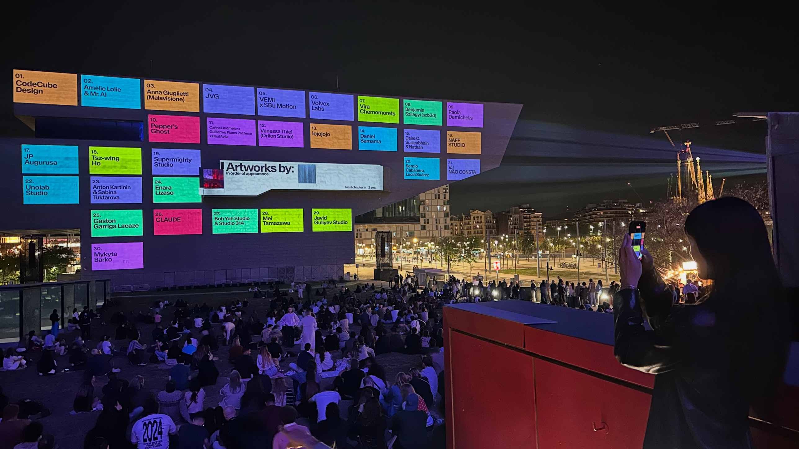 The credits wall projected on the Disseny Hub facade after the block of works finished. Thirty colored tiles with artist names, in order of appearance. Position 02, on a cyan tile, reads 'Amélie Lolie & Mr. AI'.