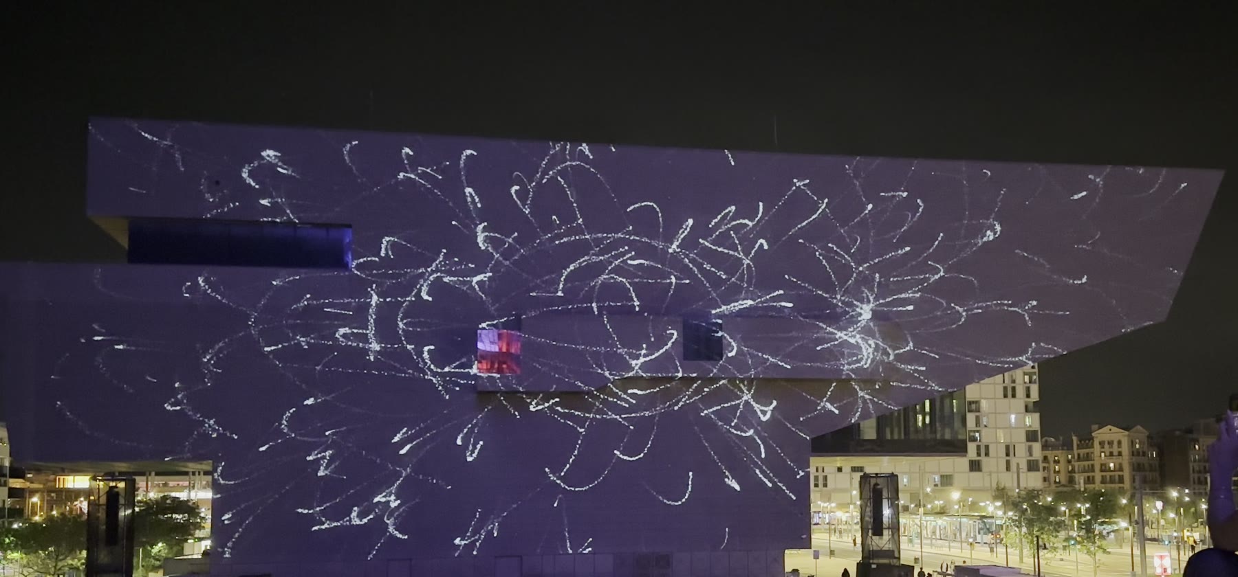 EMPREMTA — approaching the final phase. Dense radiating strokes spread outward across the facade from a central region, particles in motion just before convergence.