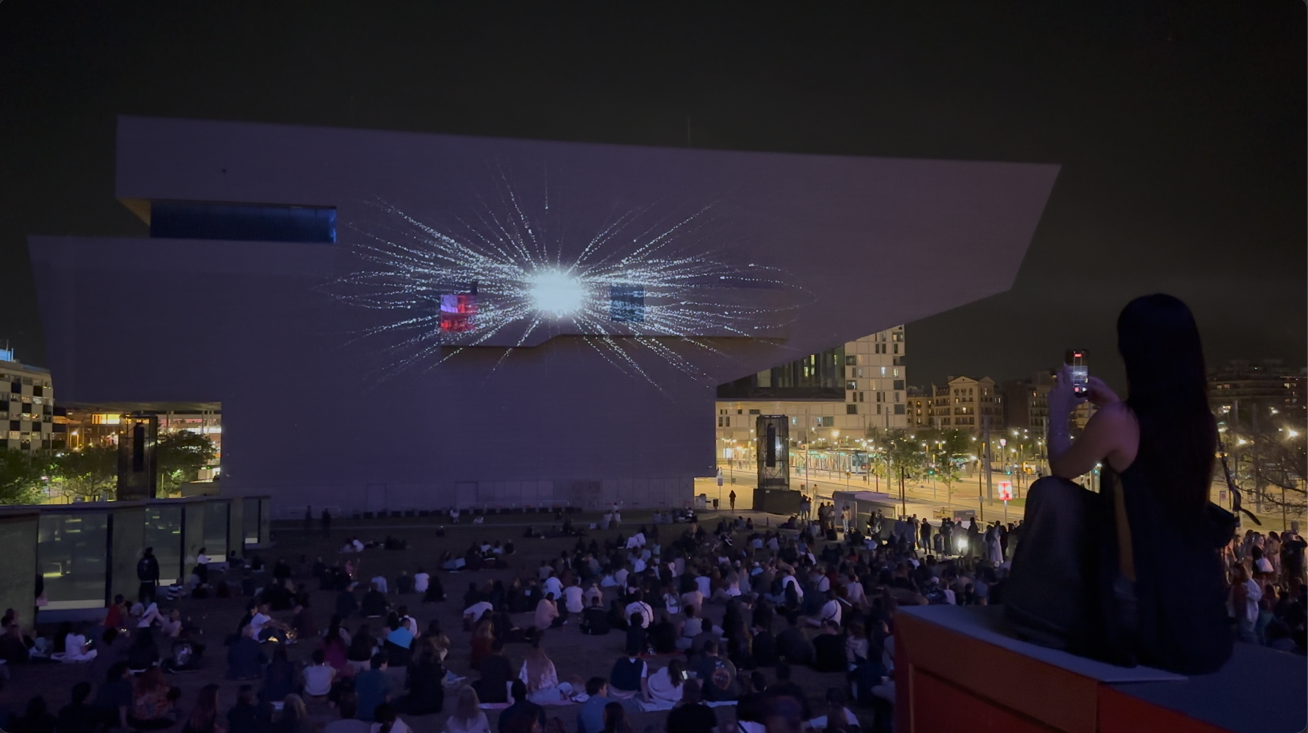 EMPREMTA — final moment. A concentrated bright point at the center of the facade with particles radiating outward in thin streaks, the last second of the piece before the facade goes dark.