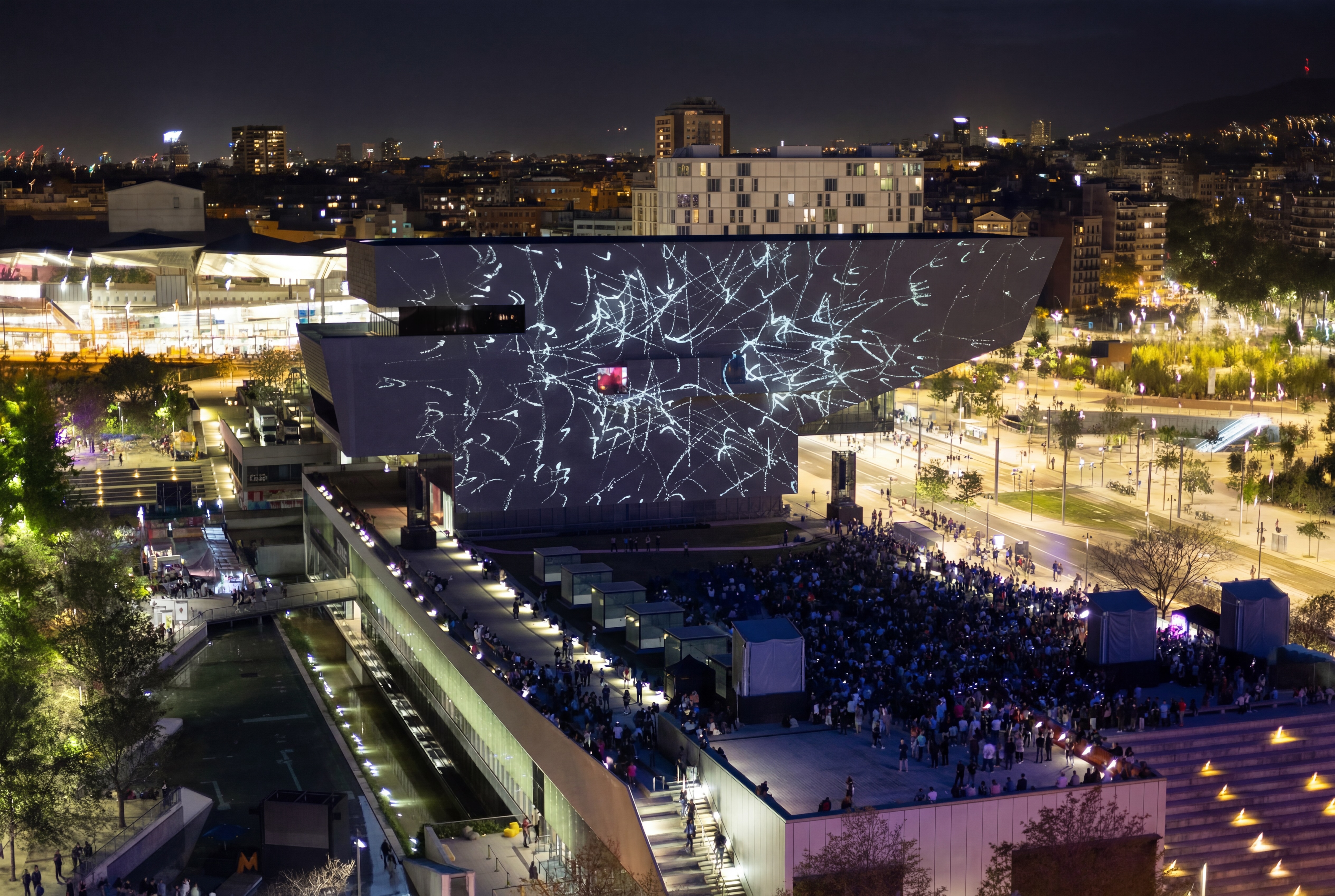 Drone shot of EMPREMTA on the Disseny Hub facade. The piece covers the dark stepped cantilever with pale handmade-looking strokes, the lit plaza and crowd below, the Barcelona cityscape extending to the horizon.