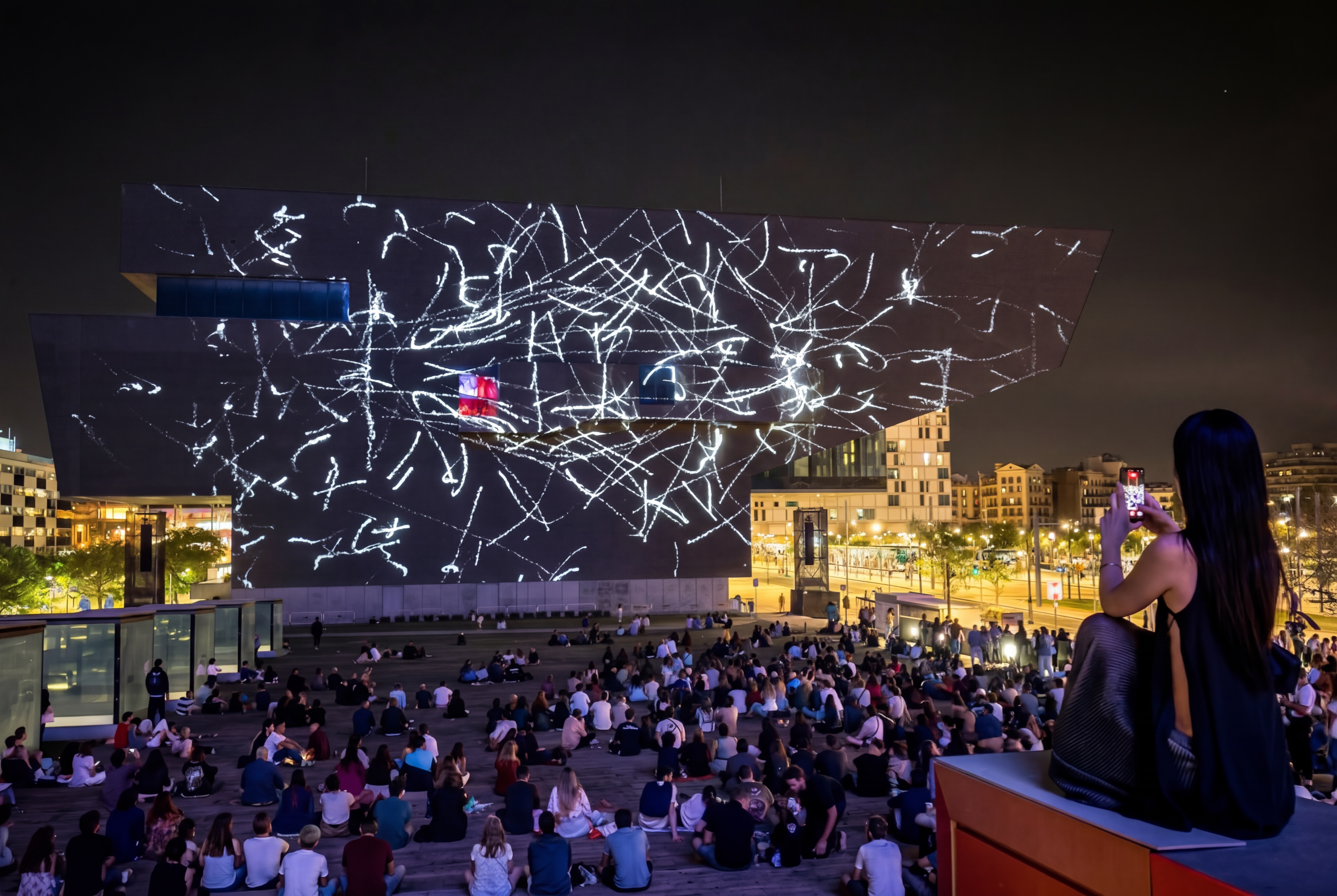Amélie Lolie at Plaça de Santiago Pey, on an elevated ledge on the right side of the plaza, holding up her phone to film EMPREMTA projected on the facade. The crowd sits on the plaza below; the facade glows with pale handmade strokes against the night sky.